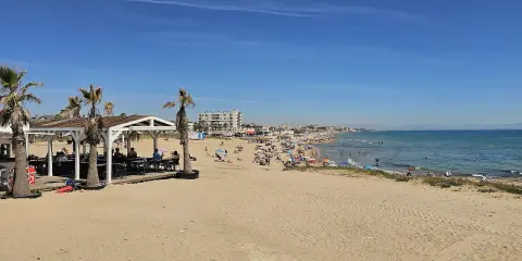 Sonniger Tag am Playa de la Mata. Menschen entspannen sich am Strand, einige im Wasser, andere unter Sonnenschirmen. Palmen säumen den Sand.