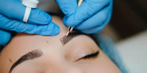 Main d'une esthéticienne en bleu, effectuant un entretien microblading sur les sourcils d'une cliente.