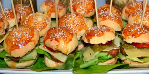 Mini hamburgery z sezamem na patyczkach, ułożone na talerzu z sałatą i dodatkami.