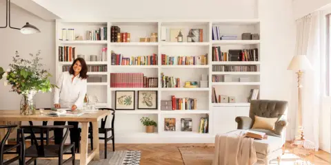 Una diseñadora casa sonríe junto a una mesa de comedor de madera, rodeada de estanterías llenas de libros y objetos decorativos.
