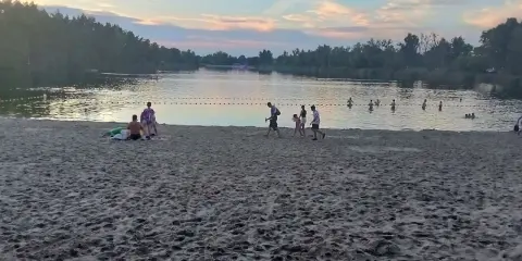 Zachód słońca nad stawem w Jelczu Laskowicach. Ludzie spacerują po piaszczystej plaży i kąpią się w wodzie.