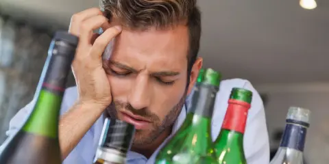 Mężczyzna z ręką na czole, otoczony butelkami, wyraźnie cierpi z powodu objawów przepicia alkoholowego.