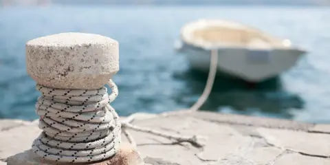 Gros plan sur un bollard en pierre avec une corde enroulée, prêt pour l'attache bateau pour quai. Un petit bateau est amarré en arrière-plan.