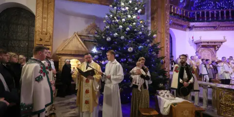 Pasterka w kościele w Zakopanem. Ksiądz czyta z księgi przy choince, obok ministranci i wierni w tradycyjnych strojach.
