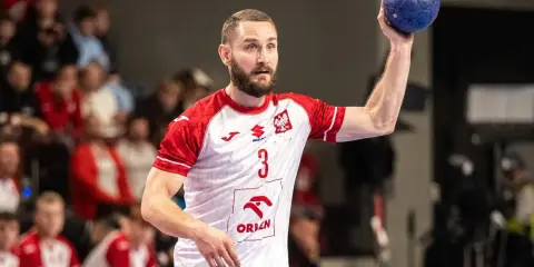Piłkarz ręczny w białej koszulce z czerwonymi rękawami, z numerem 3, przygotowuje się do rzutu karnego.