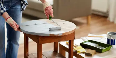 Une personne en jean et chemise à carreaux, portant des gants, utilise un rouleau pour appliquer une peinture grise sur une table ronde en bois. Le **décapage bois** est en cours pour un relooking.