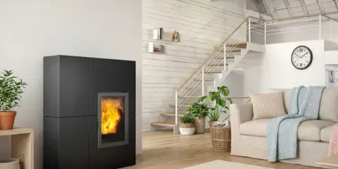 Estufa de pellets negra con fuego encendido, junto a escaleras y sofá. Ideal para un esquema de instalación de hidroestufas de pellets.