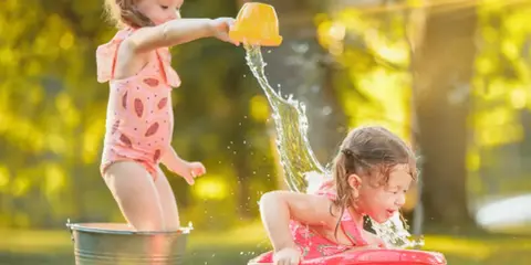 Bambine si divertono con giochi d'acqua al nido, una spruzza l'altra con un secchiello giallo.