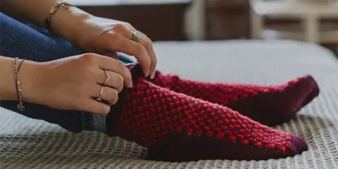 Manos ajustando calcetines rojos y morados tejidos a mano con agujas circulares.
