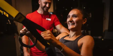 Frau lacht und trainiert mit TRX-Bändern, ihr Trainer lächelt. Sie will schnell wieder fit werden.
