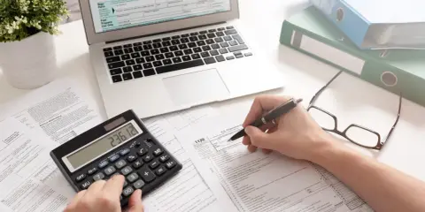 Osoba wypełnia formularz podatkowy, korzystając z kalkulatora i laptopa. To może być początek drogi, jak zostać księgowym.
