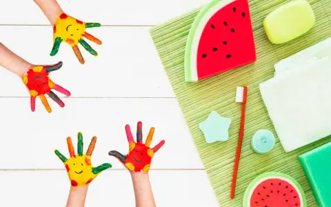 Manos pintadas de colores con caritas sonrientes, listas para hacer jabones para niños. Hay jabones con forma de sandía y otros accesorios de baño.
