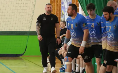 Maciej Reichel, trener, obserwuje mecz piłki ręcznej. Zawodnicy w niebieskich koszulkach czekają na swoją kolej.