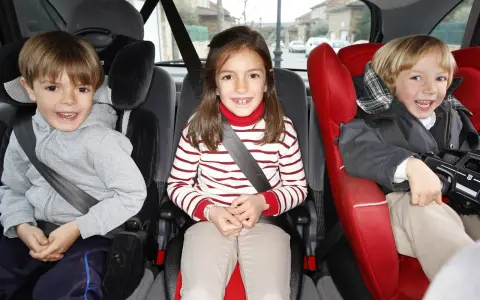Tres niños sonríen en sus asientos de coche. El niño de la izquierda, con sudadera gris, parece preguntarse cuándo puede ir un niño delante.