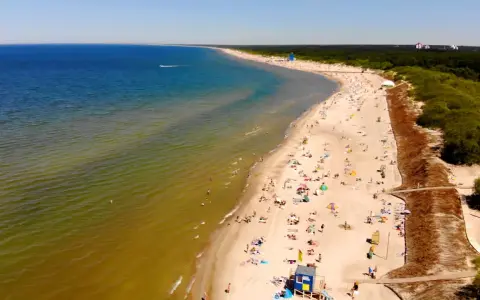 Słoneczna plaża na Litwie, gdzie turkusowe morze łagodnie przechodzi w zielonkawe wody. Tłumy plażowiczów odpoczywają na piasku, ciesząc się letnim dniem.