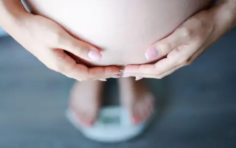 Dłonie obejmujące brzuch ciężarnej, stojącej na wadze. Schudłam w ciąży 10 kg, co widać na wadze.