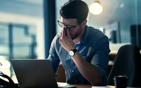 Mann reibt sich erschöpft die Augen vor dem Laptop. Ein Hackerangriff auf Privatpersonen kann zu solchen Belastungen führen.