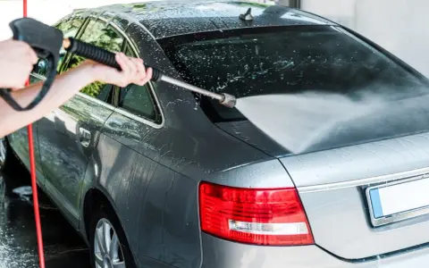 A person demonstrates how to power wash a car, spraying water onto the rear of a grey sedan.