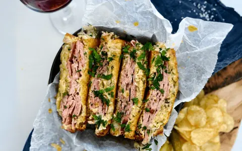 En saftig reuben sandwich, skuren i fyra bitar och toppad med färsk persilja. Serveras med krispiga potatischips och en glas rödvin.