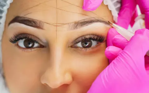 Nahaufnahme einer Frau, deren Augenbrauen mit Linien markiert werden, während eine Person in pinken Handschuhen mit einem Stift arbeitet.