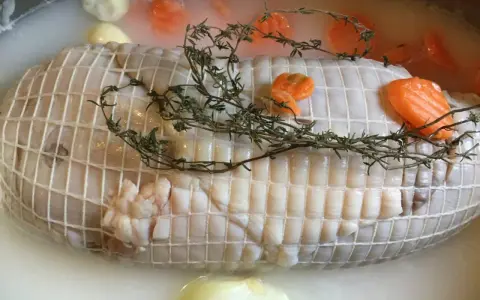 Préparation de la cuisson tête de veau cocotte. Le veau est ficelé, entouré de carottes et de thym dans un bouillon.