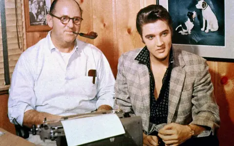 Tom Parker und Elvis Presley sitzen nebeneinander. Parker, mit einer Pfeife im Mund, tippt auf einer Schreibmaschine.
