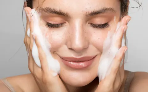 Donna con gli occhi chiusi mentre si lava il viso con un detergente schiumoso. Un gesto delicato per capire come si usa il detergente viso.