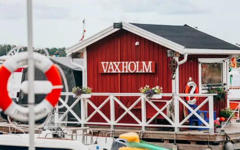 Rött hus med texten VAXHOLM vid vattnet. En livboj hänger på väggen. Vaxholms kastell skymtar i bakgrunden.