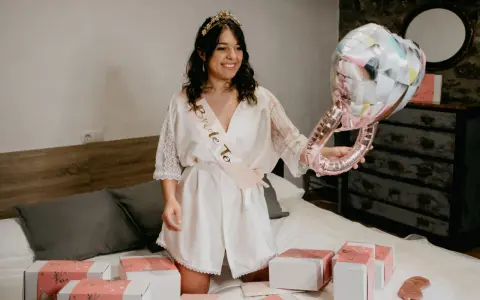 Novia sonriente con corona y banda "Bride To Be", sosteniendo un globo de anillo. Alrededor, cajas de regalo y un pack de accesorios para despedida de soltera.