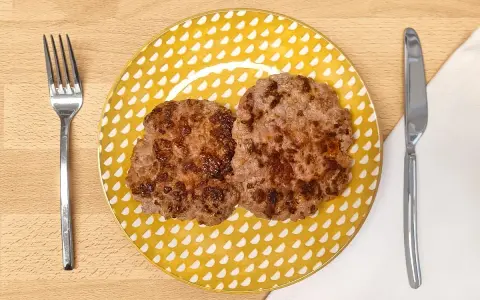 Dos hamburguesas para bebes, doradas y jugosas, servidas en un plato amarillo con puntos blancos. Cubiertos a los lados.