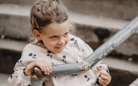 Mała dziewczynka z spiętymi włosami i spinką, w bluzie w kwiatki, opiera się o poręcz. Jej spojrzenie sugeruje dziwne zachowanie 2,5 latka.