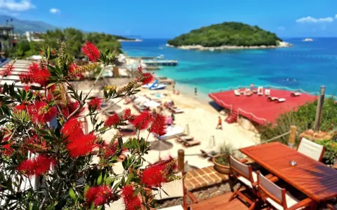 Albania camping nad morzem: czerwone kwiaty na tarasie z widokiem na piaszczystą plażę, turkusowe morze i zieloną wyspę.