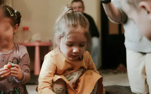 Skupiona dziewczynka w pomarańczowej bluzce, jak bawi się dziecko z autyzmem, z uwagą przygląda się swoim dłoniom.