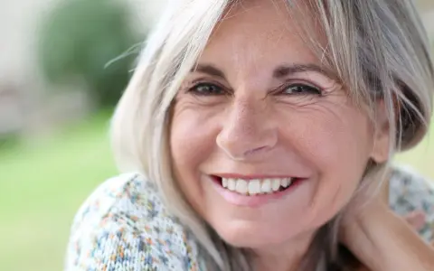 Mujer sonriente con cabello rubio y arrugas normales a los 50 años, disfrutando de un día al aire libre.