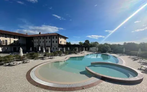 Piscina a forma di rene con idromassaggio annesso, circondata da lettini e ombrelloni, di fronte a un hotel in stile piemontese.