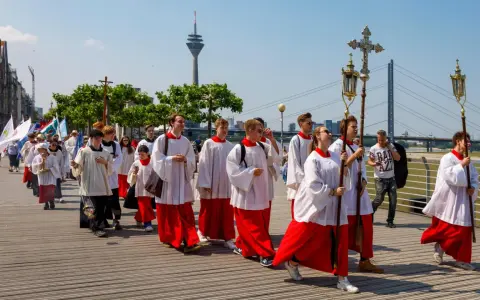 Jugendliche in Soutanen und Alba nehmen an einer Prozession teil, die an christliche Feiertage erinnert. Sie tragen Kreuze und Fahnen.