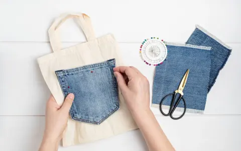 Mains épinglant une poche extérieure en jean sur un sac en toile. Ciseaux et chutes de tissu à proximité.