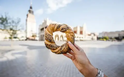 Ręka trzyma obwarzanka krakowskiego na tle Sukiennic i wieży Ratuszowej.