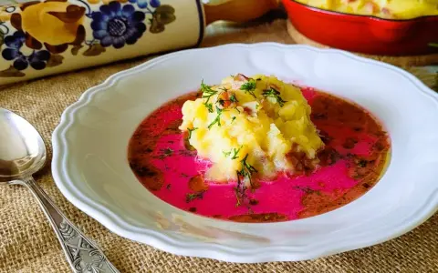 Pyszny barszcz czerwony zabielany, przepis babci. Na talerzu purée ziemniaczane z dodatkiem boczku i koperku, zanurzone w intensywnie różowym barszczu.