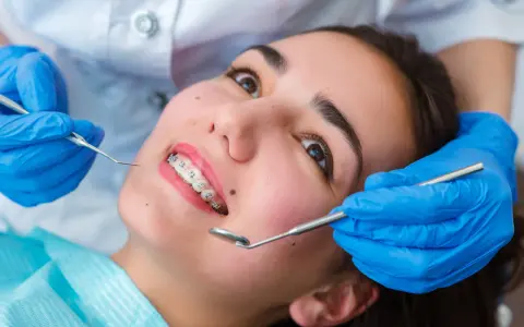 Młoda kobieta z aparatem ortodontycznym na zębach podczas wizyty u ortodonty. Dentysta w niebieskich rękawiczkach bada jej uśmiech.
