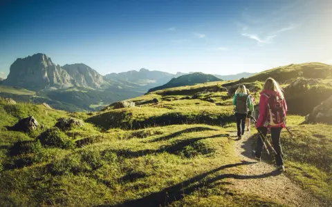 Persone in un percorso di trekking e hiking tra le montagne, con zaini e bastoncini da passeggio, sotto un cielo azzurro.