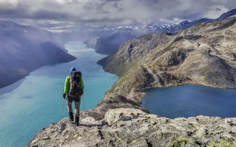 Vandrare med ryggsäck står på en klippa och blickar ut över turkosa sjöar och berg i Jotunheimen nationalpark.