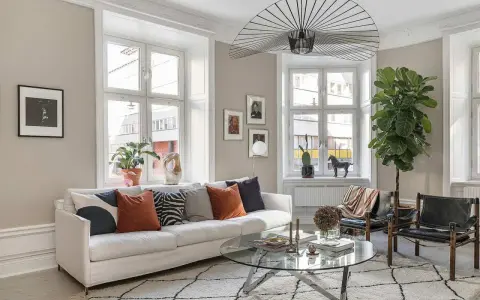Vardagsrummet är ljust med en vit soffa, kuddar i olika färger och mönster, ett runt soffbord och en stor taklampa. En stor grön växt bidrar till trivseln.