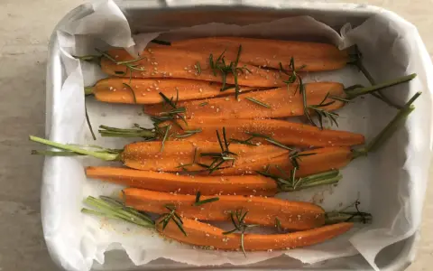 Carote al forno, tagliate a metà, condite con rosmarino e semi di sesamo, pronte per essere infornate.