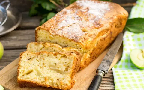 Szybkie ciasto z jabłkami, posypane cukrem pudrem, pokrojone na kromki, leży na desce obok jabłek i noża.