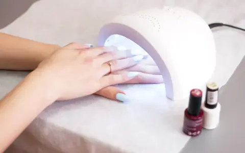 Dłonie z jasnoniebieskimi paznokciami pod lampą UV. W tle stoją buteleczki z lakierami. Lampa UV w ciąży - delikatna pielęgnacja.