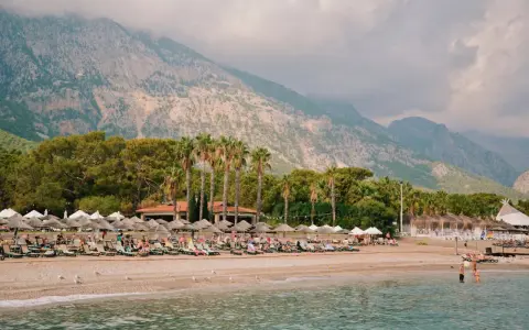Plaża w Turcji z leżakami, palmami i górami w tle. Ludzie odpoczywają, a w wodzie kąpie się kilka osób.