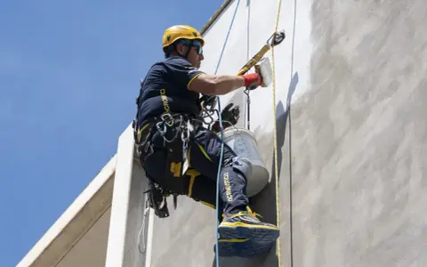 Trabajador en altura realiza instalación SATE, aplicando revestimiento en fachada con equipo de seguridad.