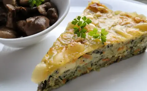 Kawałek zapiekanki z cukinii z warzywami i serem, podany z miseczką pieczarek.