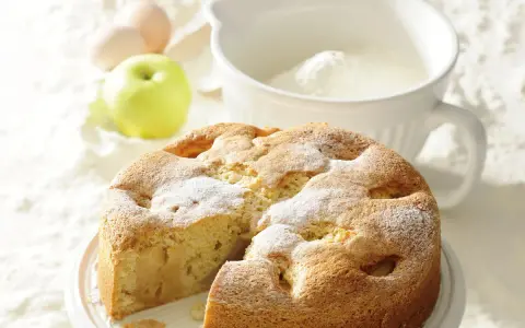 Szybkie i tanie ciasto na niedzielę: puszysty jabłecznik z cukrem pudrem, obok jabłko, jajka i miska z mąką.